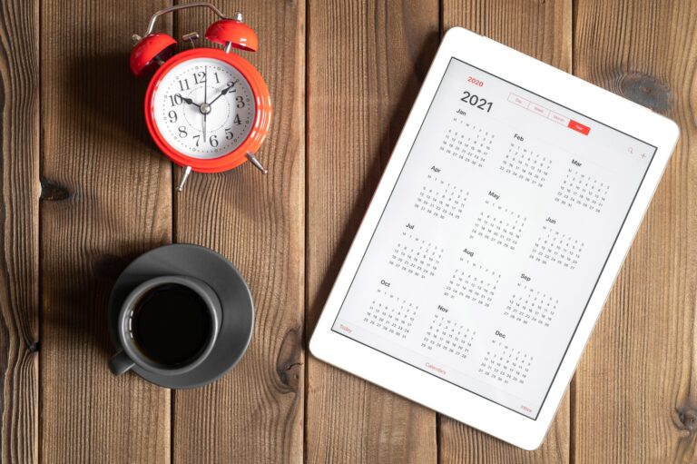 tablet with calendar for 2021, a cup of coffee and a red alarm clock on a wooden background flat lay