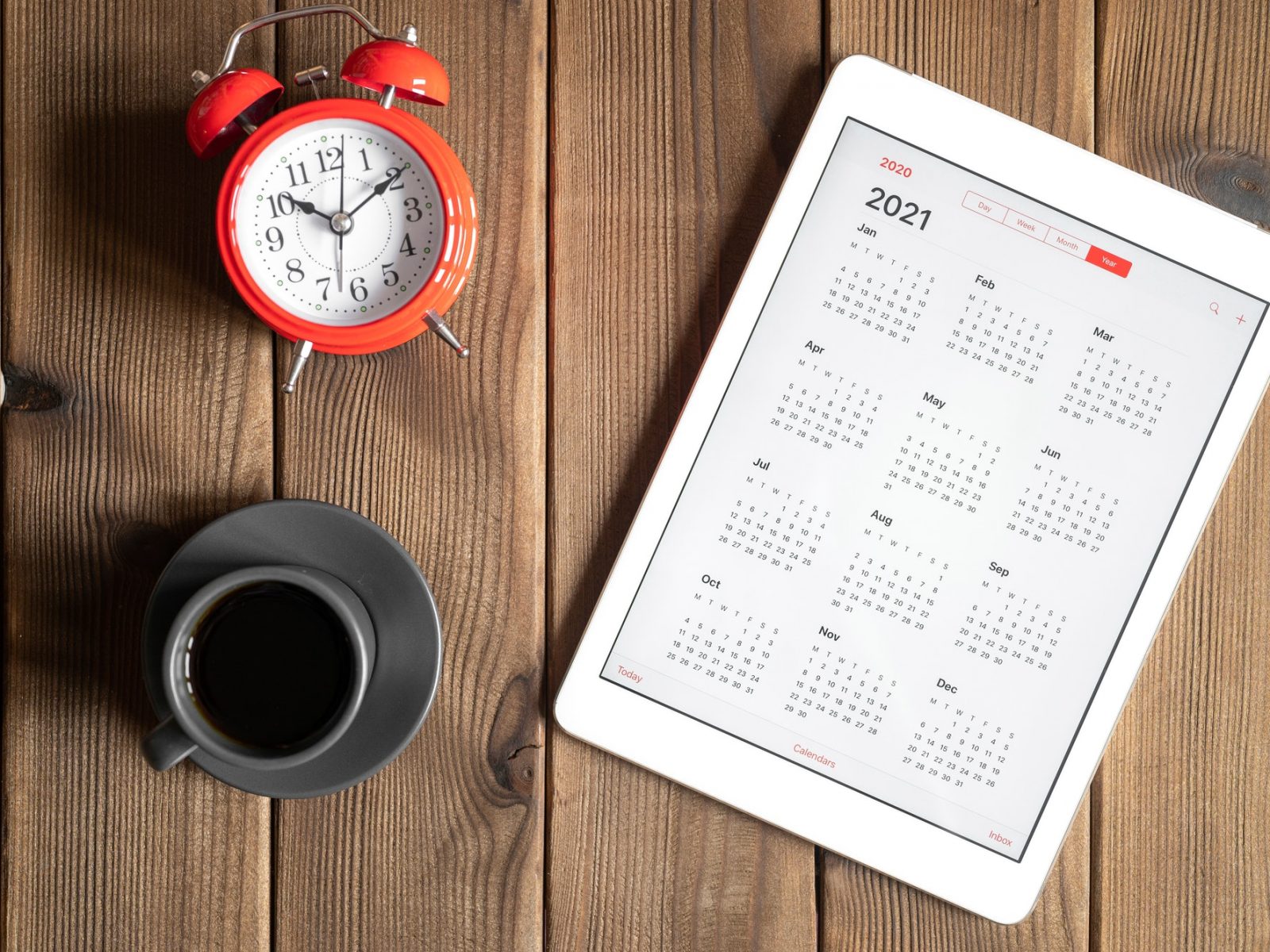 tablet with calendar for 2021, a cup of coffee and a red alarm clock on a wooden background flat lay